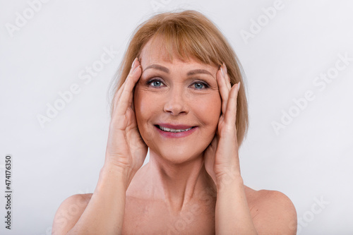 Portrait of feminine nude senior woman touching smooth skin on her face, smiling at camera on light studio background