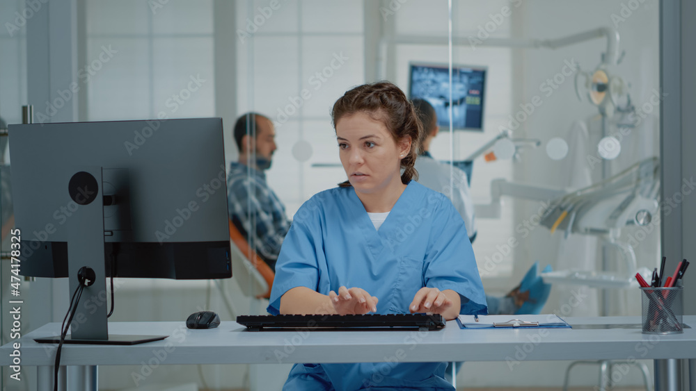 Dental assistant using computer and x ray scans at desk while stomatologist examining patient teethcare. Caucasian staff of orthodontists working at oral clinic for stomatology care Dental assistant using computer and x ray scans at desk while stomatologist examining patient teethcare. Caucasian staff of orthodontists working at oral clinic for stomatology care
