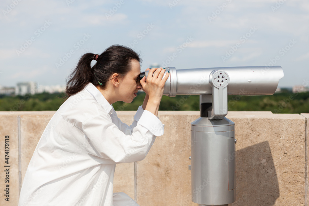Beautiful woman looking through tourist binocular telescope observing panoramic view of metropolitan city. Caucasian female standing at tower observation point. Landscape with urban buildings