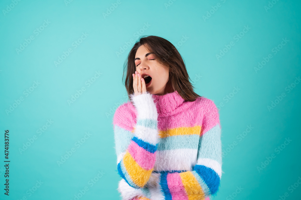Young woman in a bright multicolored sweater on a blue background sleepy eyes, cute yawns, lack of sleep