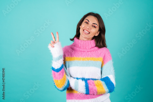 Young woman in a bright multicolored sweater on a blue background cute smiling  playful  showing tongue and v gesture