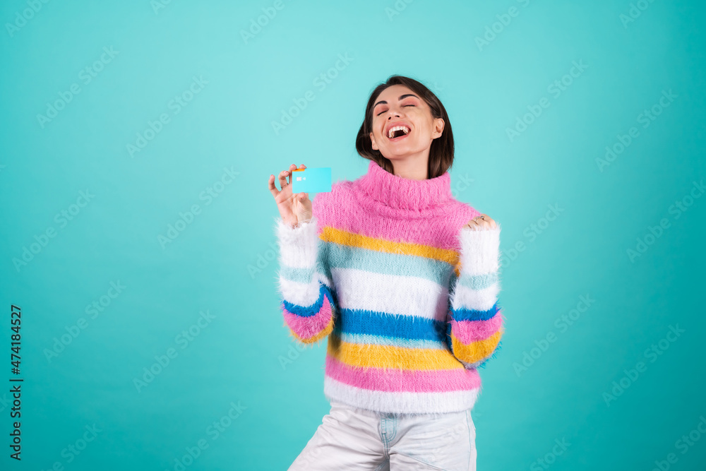 A young woman in a bright multicolored sweater on a blue background shows a credit card, jumps with happiness, is very happy, a winner gesture A young woman in a bright multicolored sweater on a blue background shows a credit card, jumps with happiness, is very happy, a winner gesture