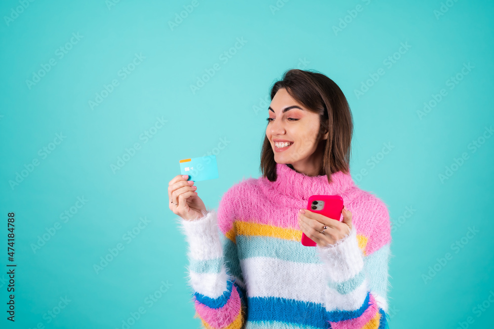 Young woman in a bright multicolored sweater on a blue background holds a credit card, pays for purchases shopping online on a mobile phone, joyful smiles Young woman in a bright multicolored sweater on a blue background holds a credit card, pays for purchases shopping online on a mobile phone, joyful smiles