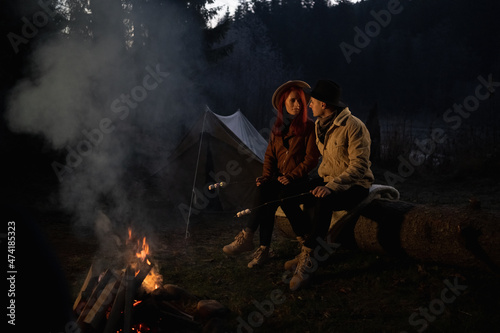 A couple in love, a man and a woman sit at night in the woods by the fire near the tent, holding sticks with marshmallows in their hands. The concept of hiking and recreation in the forest.
