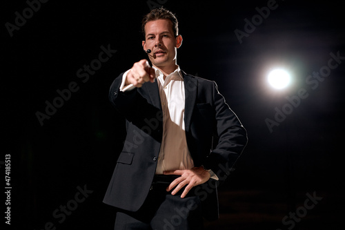 speaker with microphone pointing finger to the listener, standing on stage with projector in background, concepts of speakers and communication. American male in formal suit giving speech