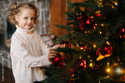 Portrait of smiling little blonde curly child girl decorating Christmas tree with festive balls and toys in cozy living room looking at camera. Cute kid preparation home to holiday celebration.