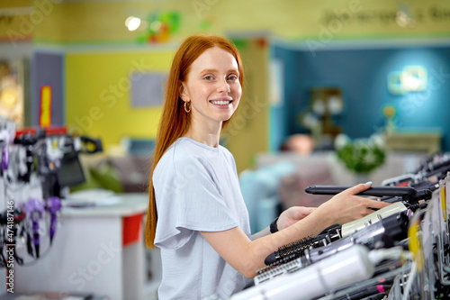 Smiling redhead woman with long hair  buying curling iron in store. Female is standing by shelves with household appliances. The concept of buying household appliances for beauty and self-care