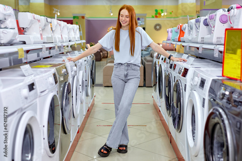 Woman buying new clothes washer in supermarket, choosing the best and the most modern. Redhead Lady In Casual Wear Standing Between Various Different Washine Machines, Make Choice and purchase photo