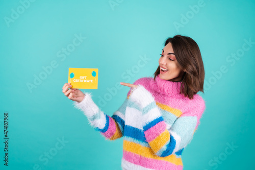 Young woman in a bright multicolored sweater on a blue background holds a gift certificate  smiles enthusiastically  beautiful makeup