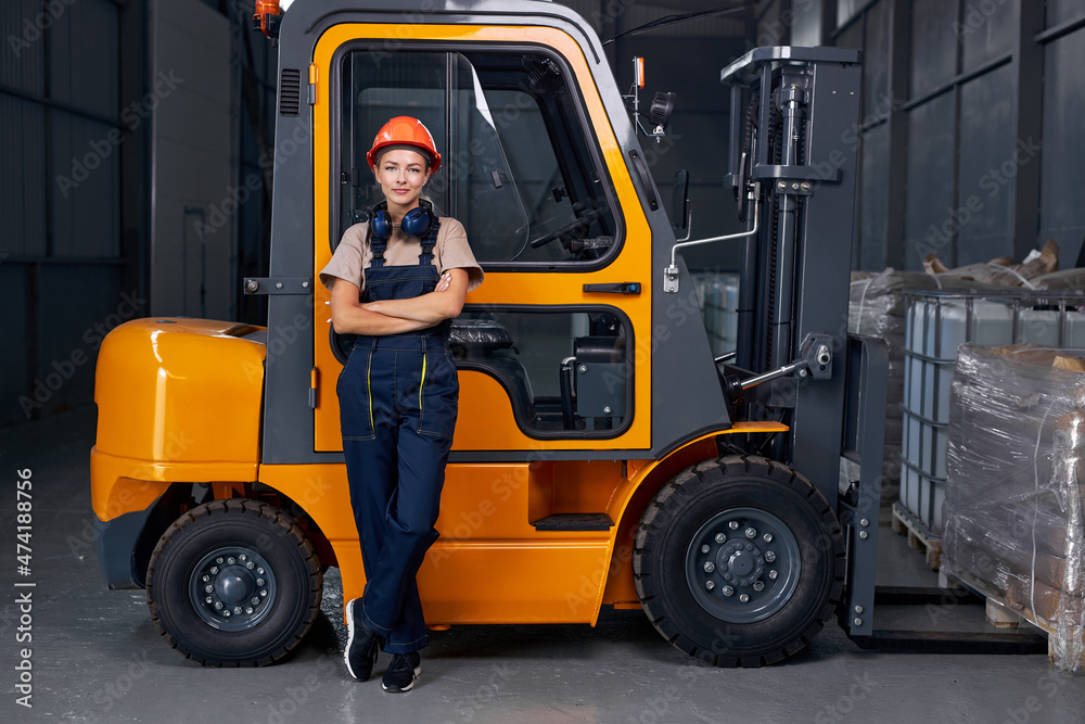 smiling woman labor worker near forklift driver in industry factory logistic shipping warehouse, posing with arms folded, dressed in uniform and orange protective hardhat, after successful good job