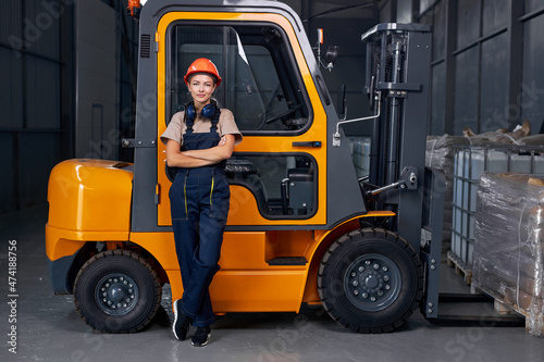 smiling woman labor worker near forklift driver in industry factory logistic shipping warehouse, posing with arms folded, dressed in uniform and orange protective hardhat, after successful good job