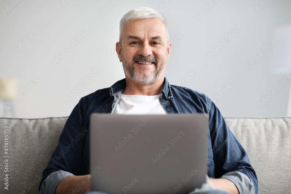 Cheerful mature man using laptop while resting on sofa Cheerful mature man using laptop while resting on sofa