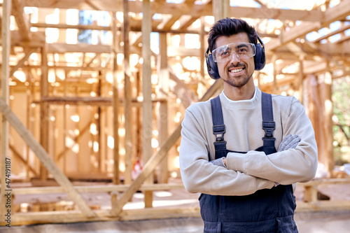smiling caucasian man in headset and uniform at work place, wooden house. Bearded young male in protective spectacles look at camera holding arms folded. architecture, renovation, construction