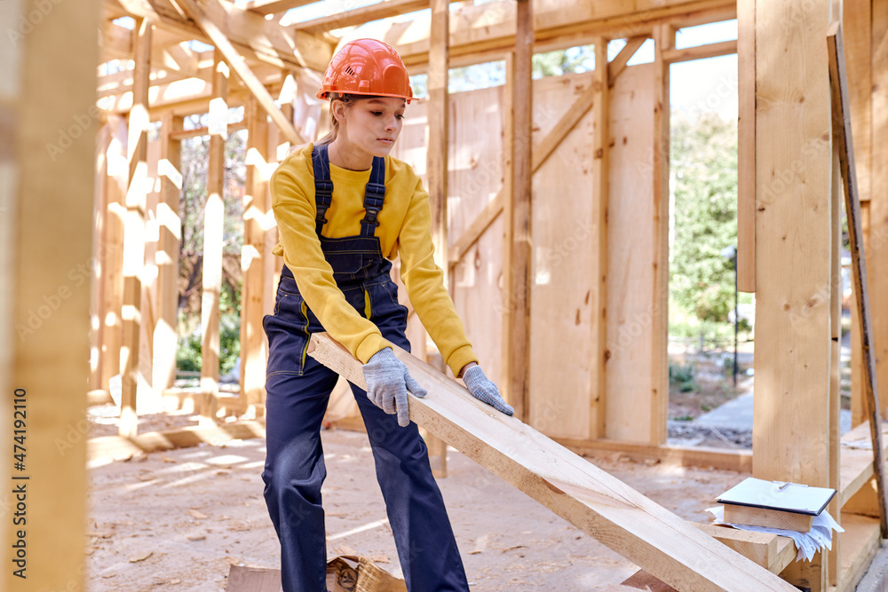 Confident female constructor wearing helmet and uniform while preparing frame for covering roof on the future house, dragging wooden beam into another place, concentrated on work Confident female constructor wearing helmet and uniform while preparing frame for covering roof on the future house, dragging wooden beam into another place, concentrated on work