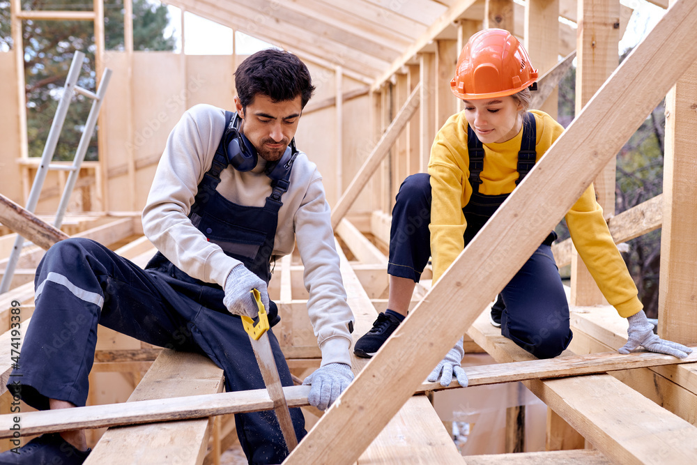 Caucasian Contractors Carpenters Workers Using Wood Saw in Construction Zone. Wooden Skeleton Framing Building. Hardworking Cooperative Man And Woman In Uniform Concentrated On Work Caucasian Contractors Carpenters Workers Using Wood Saw in Construction Zone. Wooden Skeleton Framing Building. Hardworking Cooperative Man And Woman In Uniform Concentrated On Work