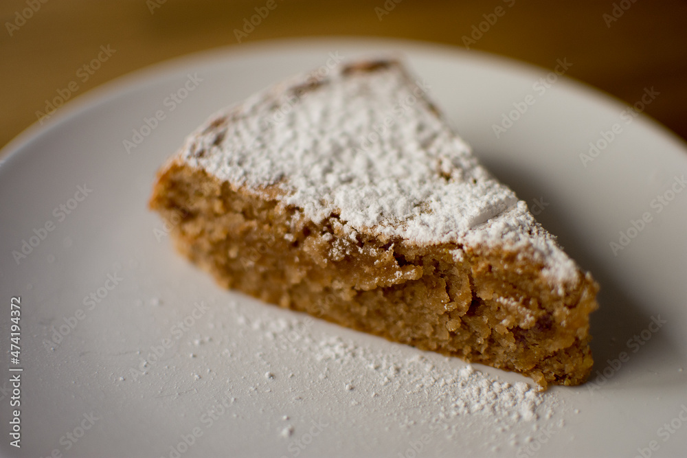 Piece of homemade Santiago cake covered with glass sugar in a white plate on a wooden table, with glass sugar on the plate. Front view copy space.