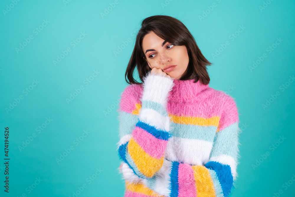 A young woman in a bright multicolored sweater on a blue background stands with a bored indifferent look, a fist on her chin A young woman in a bright multicolored sweater on a blue background stands with a bored indifferent look, a fist on her chin