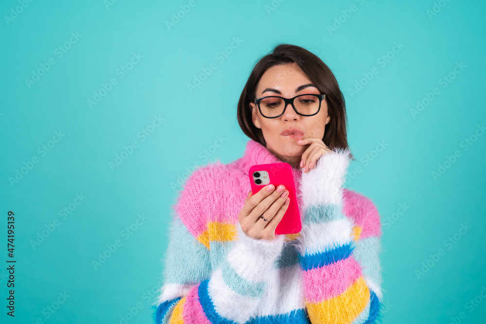 Young woman in a bright multicolored sweater on a blue background with glasses, holds the phone, pensive, dreamy looks at the screen, bites her lip Young woman in a bright multicolored sweater on a blue background with glasses, holds the phone, pensive, dreamy looks at the screen, bites her lip