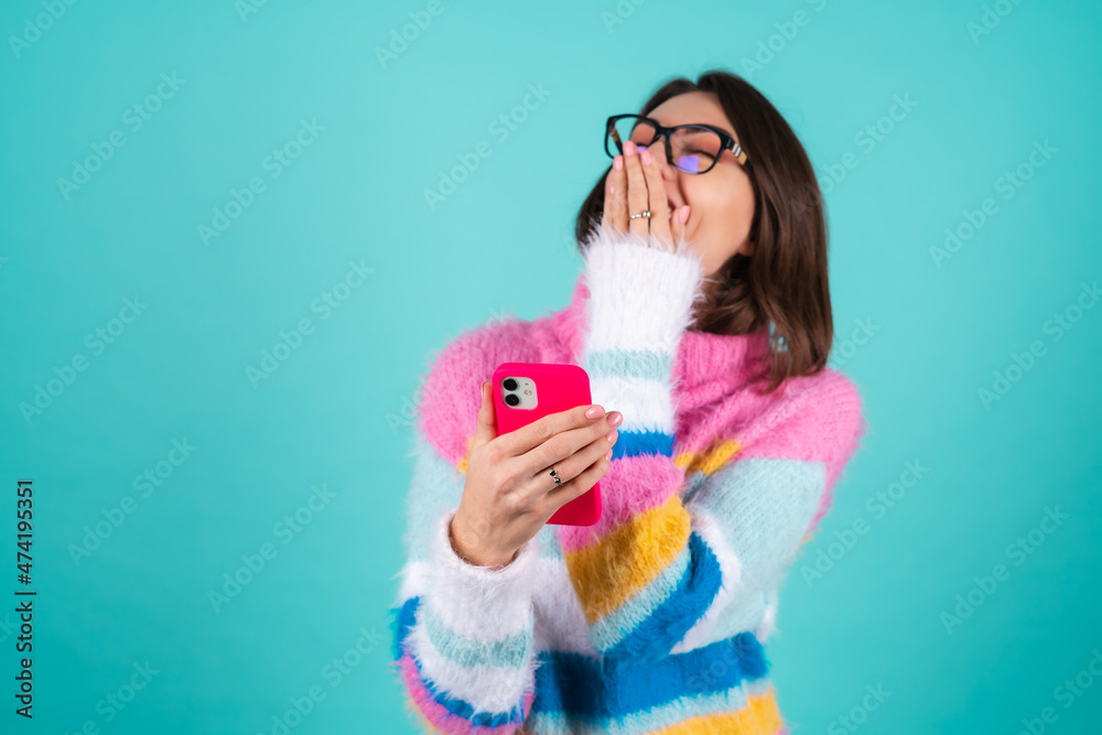 A young woman in a bright multicolored sweater on a blue background with glasses, holds the phone, laughs cheerfully, read a joke, anecdote, covers her mouth with her hand, laughs nonstop A young woman in a bright multicolored sweater on a blue background with glasses, holds the phone, laughs cheerfully, read a joke, anecdote, covers her mouth with her hand, laughs nonstop