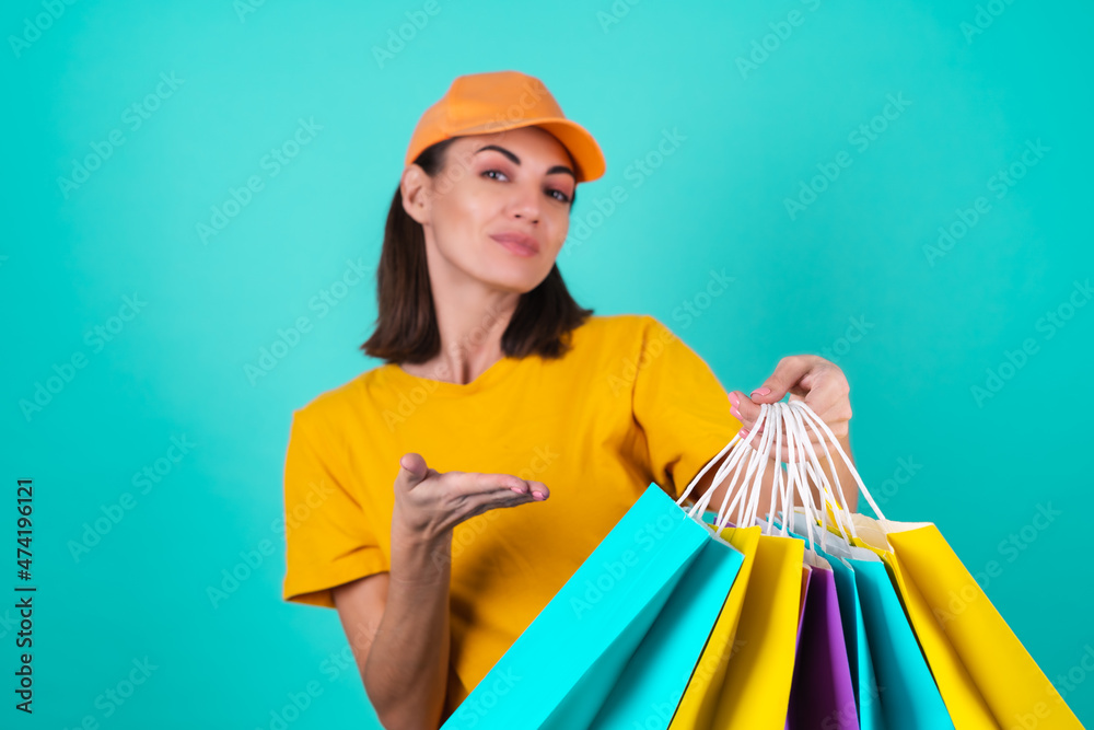 Young woman delivery service worker, courier, in an orange yellow cap on a blue background, holds multicolored shopping paper bags, delivers an order Young woman delivery service worker, courier, in an orange yellow cap on a blue background, holds multicolored shopping paper bags, delivers an order