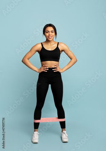 Fitness equipment. Smiling black woman in sportswear doing exercises with elastic band, isolated on blue background