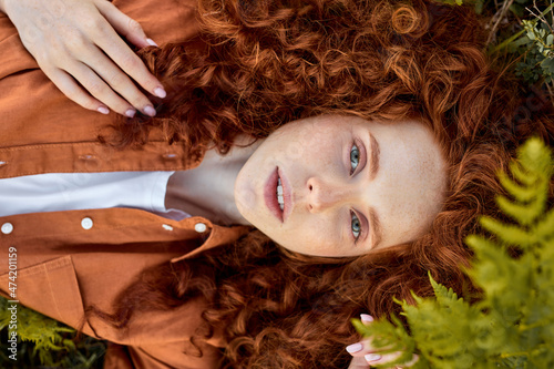 Fotografía Top view on redhead curly female of caucasian appearance lying on grass in summer, posing looking at camera