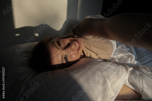 Young blond woman taking a smiling selfie on her bed with a white cover, newly awakened woman taking selfie