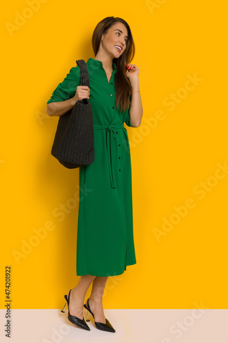 Beautiful young woman in long button-up green dress is standing with black straw bag on her shoulder.