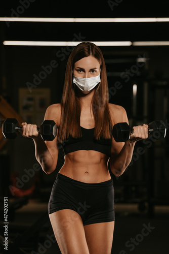 A fit woman in a face mask is doing bicep curls with dumbbells in a gym.