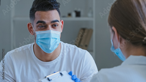 Arabic man hispanic guy patient sick male wears medical protective mask on face talks about symptoms of health problems to unrecognizable doctor woman nurse writing down notes, consultation in clinic.