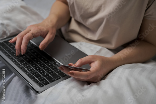 woman browsing social media on laptop in bed, woman shopping with credit card and laptop on bed with white cover, shopping online in bed