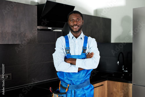 Smiling Handsome Young African Handyman Plumber In Blue Uniform. Young Skilled Handyman In Overall Standing At Home In Kitchen Posing Looking At Camera. Repairman Service, Hard Work Concept