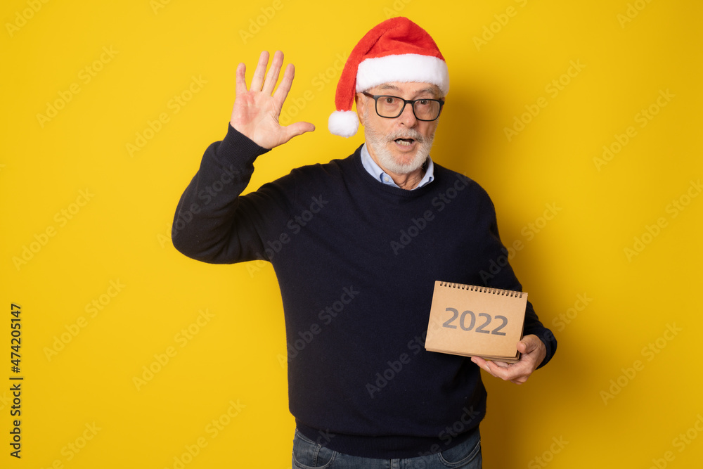 Senior excited man wearing santa hat showing calendar isolated over yellow background.