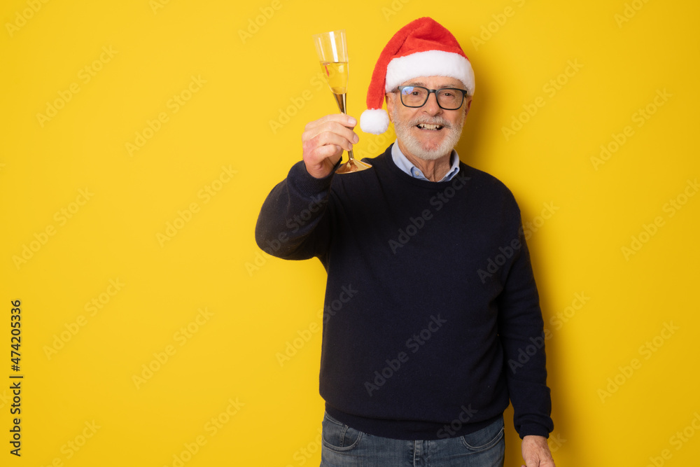 Senior man in santa hat drinking champagne standing isolated over yellow background.