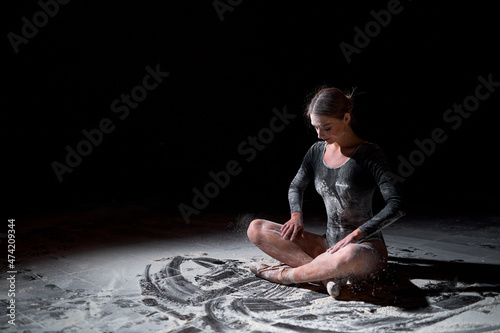 caucasian female in black bodysuit sitting on floor with crossed legs having rest. female adult ballet dancer performer in flour. Portrait of lady dancer with hair in bun sit alone isolated on black