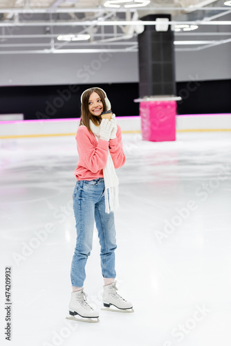 full length of happy woman in knitted sweater, ear muffs and winter outfit skating with paper cup on ice rink.