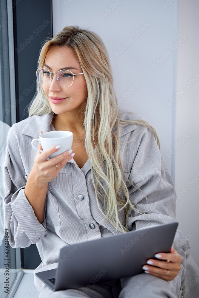 nice caucasian female in casual domestic clothing using computer laptop and sitting on window sill alone at home, drinking coffee. Freelance working, free time and leisure, online browsing internet nice caucasian female in casual domestic clothing using computer laptop and sitting on window sill alone at home, drinking coffee. Freelance working, free time and leisure, online browsing internet
