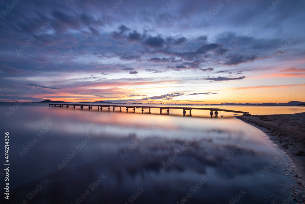 Obraz premium Photograph of a sunrise in the Mar Menor, in the region of Murcia, Spain. There is a jetty over the calm waters of the sea and colorful clouds reflecting in the water