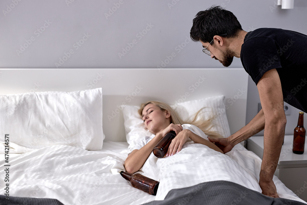 Domestic scene at home bedroom of young family husband and wife. Sleeping Lazy Drunk Blonde Woman Lying on bed while husband touching trying to wake her up. Alcohol addiction concept. focus on man Domestic scene at home bedroom of young family husband and wife. Sleeping Lazy Drunk Blonde Woman Lying on bed while husband touching trying to wake her up. Alcohol addiction concept. focus on man