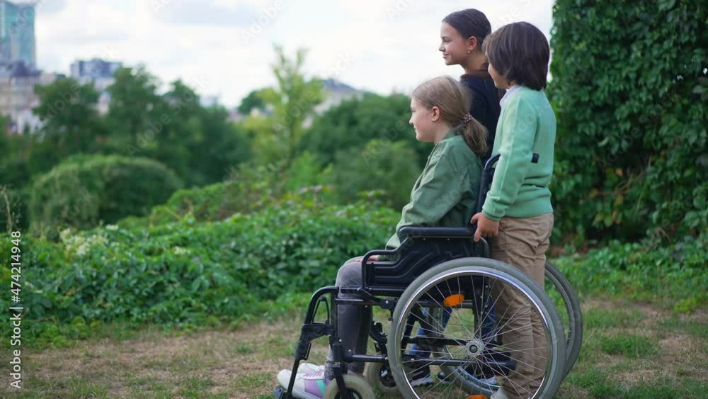 Side view wide shot happy children teenagers standing in summer park looking away at urban city. Carefree Caucasian disabled girl in wheelchair enjoying leisure with friends outdoors. Slow motion Side view wide shot happy children teenagers standing in summer park looking away at urban city. Carefree Caucasian disabled girl in wheelchair enjoying leisure with friends outdoors. Slow motion