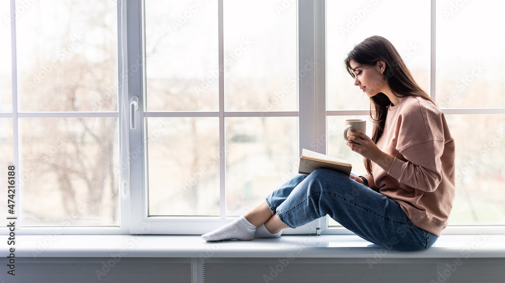 Woman sitting on windowsill drinking coffee reading book Woman sitting on windowsill drinking coffee reading book