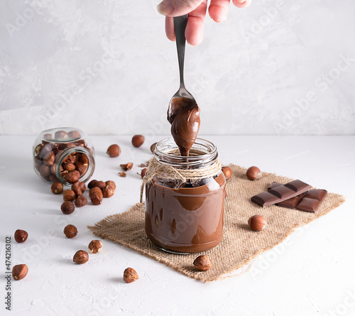 Chocolate-nut paste in a glass jar on a light table. With a spoon, the paste pours into the jar. Hazelnuts and chocolate pieces in the background photo