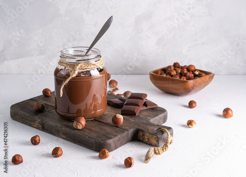 Chocolate-nut paste in a glass jar on a dark brown wooden board. Hazelnuts in a wooden bowl in the background. Nuts are scattered on the table