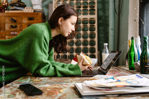 Young woman eating sandwich and using laptop in her studio