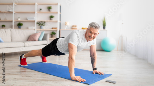 Sporty middle-aged man working out at home, standing in plank and noting time on smartphone