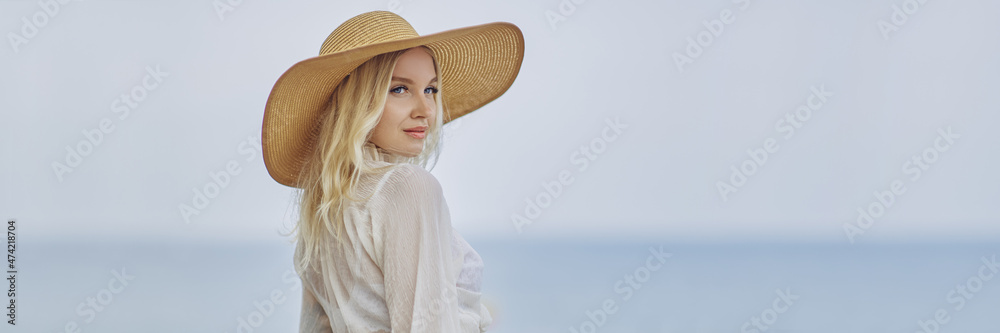 Stunning blonde woman in summer beach outfit relaxing outdoors against sea background. A fashionable romantic young adult lady wearing a trendy vintage straw hat, white blouse, and skirt, standing at