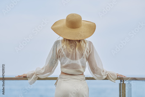 Stunning blonde woman in summer beach outfit relaxing outdoors against sea background. Back view of fashionable romantic young adult lady wearing a trendy vintage straw hat, white blouse, and skirt photo