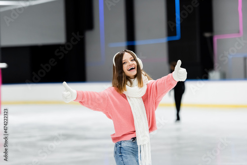 amazed young woman in pink sweater showing thumbs up on ice rink.