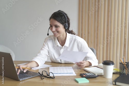 Happy woman wears headphones while sits at desk at home office looks at laptop screen works online