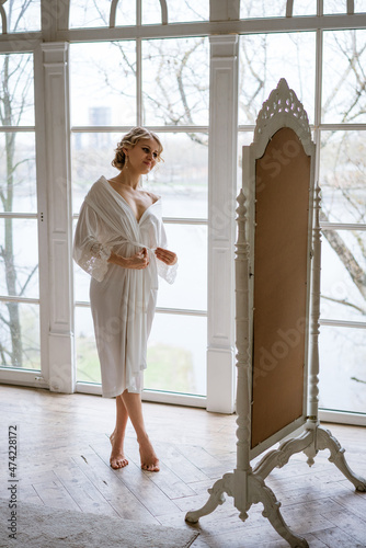 Beautiful woman in lace robe looks at her reflection in mirror while standing at home near large window. Female beauty and self-confidence. Makeup lessons. Psychological trainings. Morning of bride.